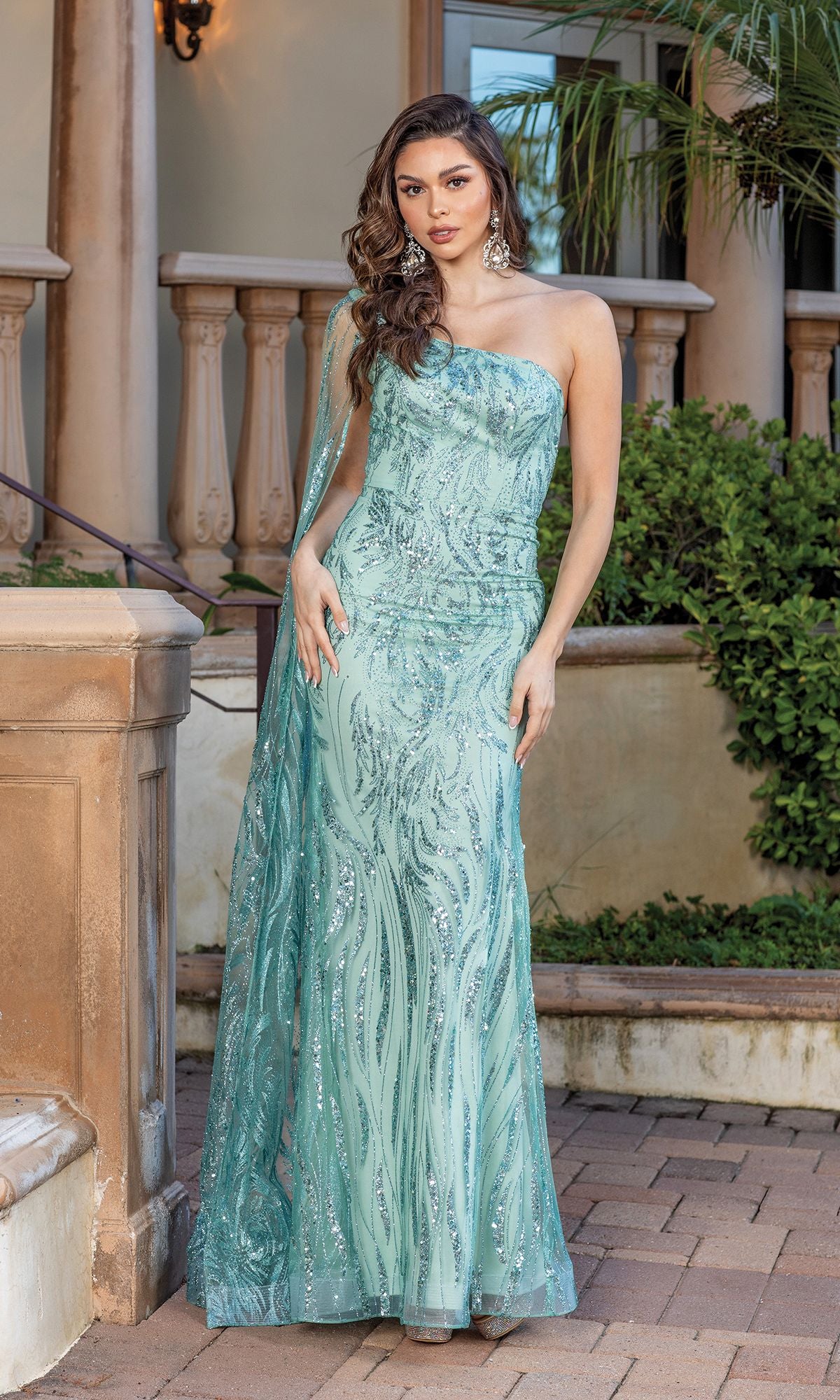 A woman stands outdoors in front of columns and greenery, wearing the One-Shoulder Long Glitter Prom Gown with Cape in light blue-green, featuring a fitted silhouette and sparkling sequin details.